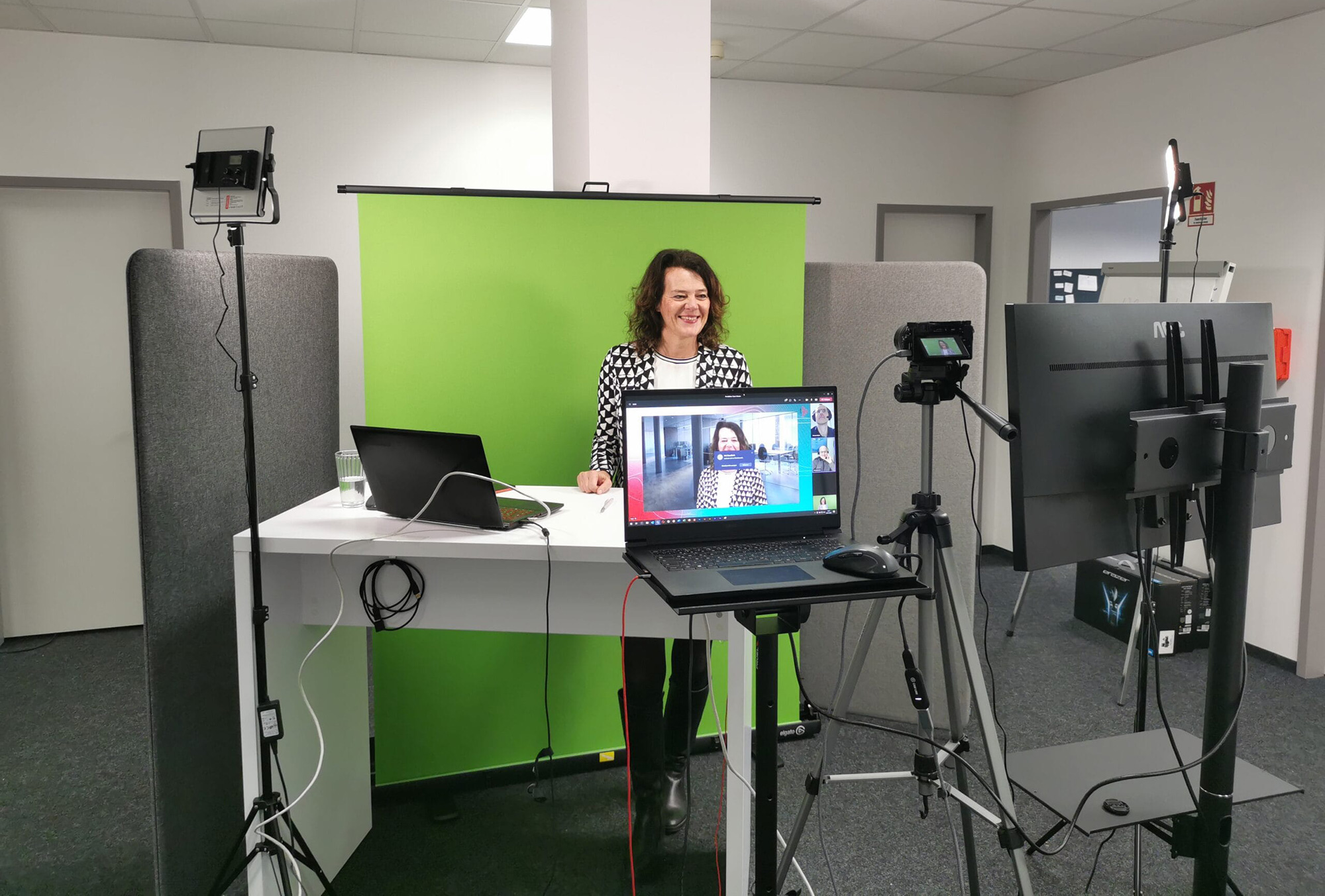Eine Frau steht vor einem Greenscreen in einem professionellen Video-Setup mit Laptop, Kamera und Beleuchtung, während sie eine Online-Präsentation hält.
